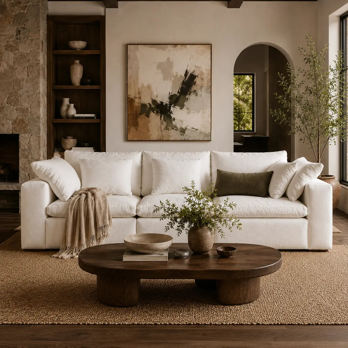 A styled living room featuring a large white upholstered sofa with cream and olive pillows, a round dark wood coffee table, a beige woven rug, and a large abstract painting on the wall.