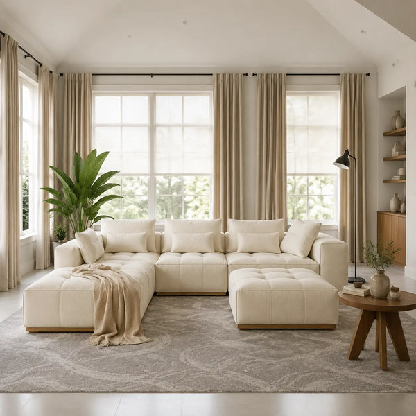 A cream tufted sectional sofa with matching pillows and a chaise ottoman in a bright, modern living room with beige curtains, a large green plant, and a round wooden side table.