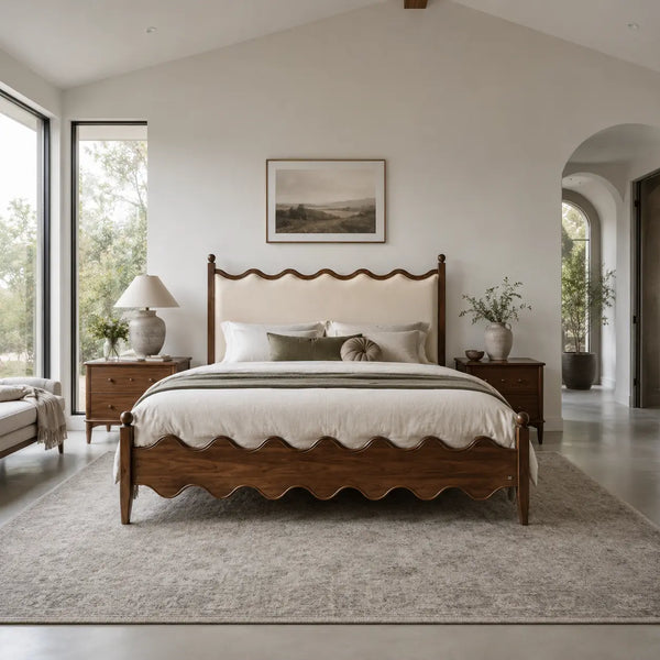 A scalloped wooden bed frame with a cream upholstered headboard is centered in a bright, modern bedroom with matching wood nightstands, a gray rug, and large windows.